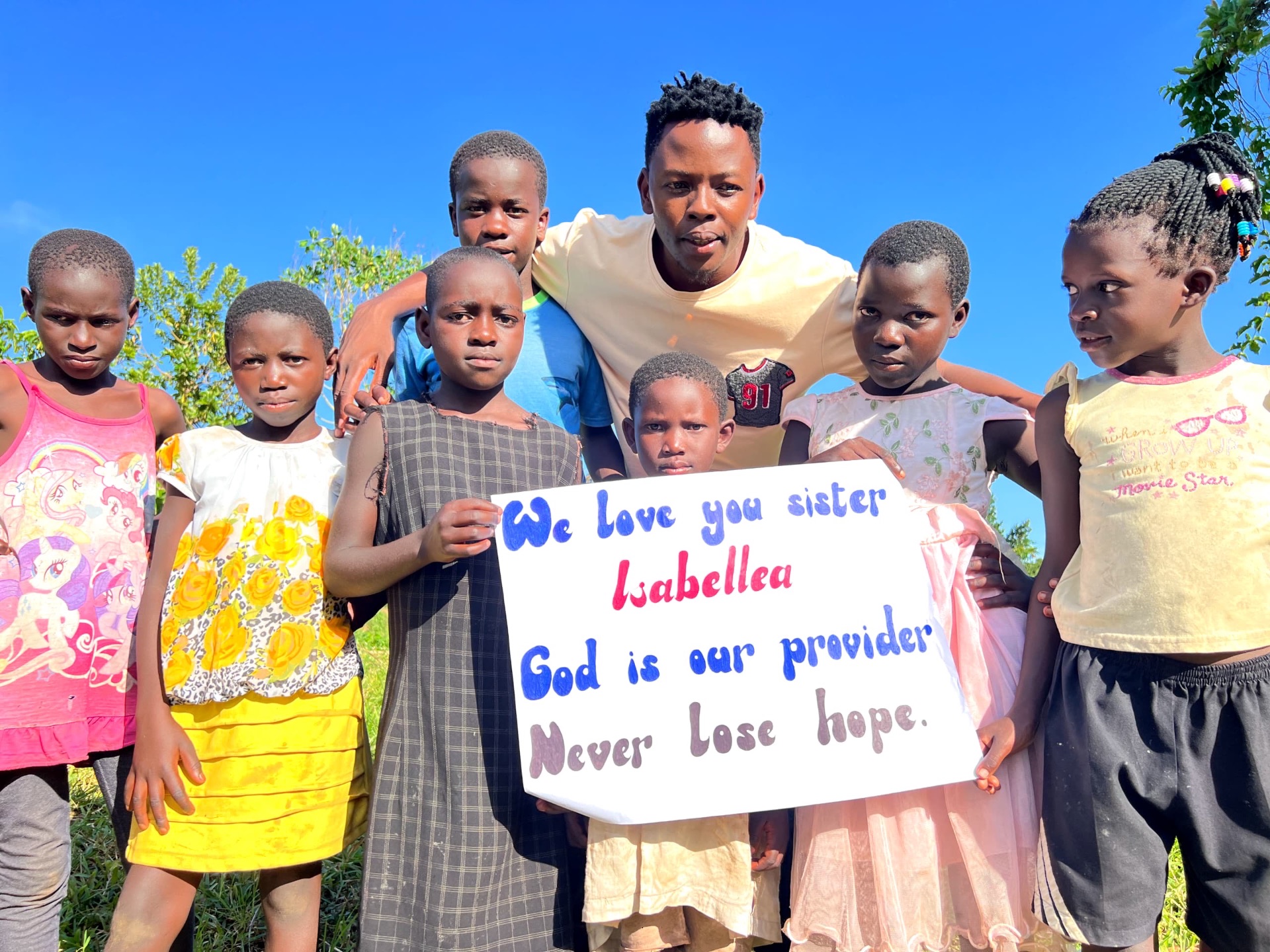 Children and staff holding a sign reading 'We love you sister Isabella — God is our provider, never lose hope.'